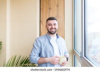 bearded handsome man stands by window with a cup in his hand on the balcony and drinks a hot coffee or tea. male in casual clothes enjoys view through in office or home, relaxing or timeout at work