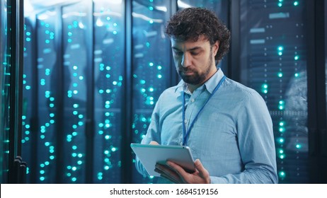 Bearded Head Administrator Running Backups On Digital Tablet In Rack Server Room. His Young Co-worker Using Laptop Updating Supercomputer Hardware. Data Center. Collaboration.