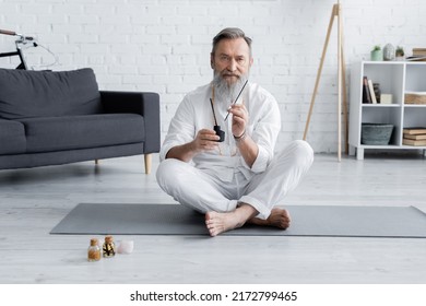 Bearded Healing Guru Sitting In Easy Pose With Aroma Sticks Near Bottles With Flavored Oils