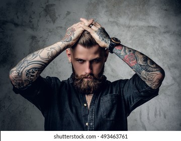 Bearded hipster male with tattoo on arms holding hands on his head.