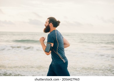 Bearded hipster man is running on the beach near sea or ocean on a cloudy day. He is wearing a phone holder on his arm.