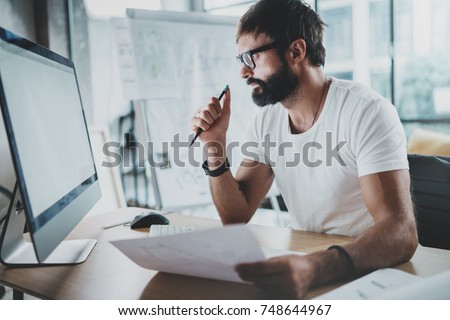Bearded hipster professional wearing eye glasses working at modern loft studio-office with desktop computer.White blank display screen.Blurred background. Horizontal