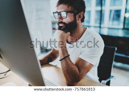 Bearded hipster professional wearing eye glasses working at modern loft studio-office with desktop computer.Panoramic windows on blurred background. Horizontal