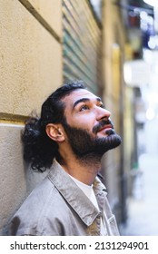 Bearded Hispanic Male In Casual Clothes With Ponytail Looking Up And Dreaming While Leaning On Wall On City Street