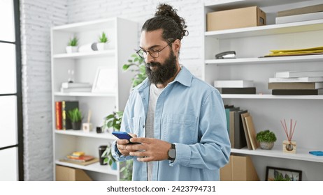 A bearded hispanic man in casual attire focused on his smartphone in a bright modern office. - Powered by Shutterstock