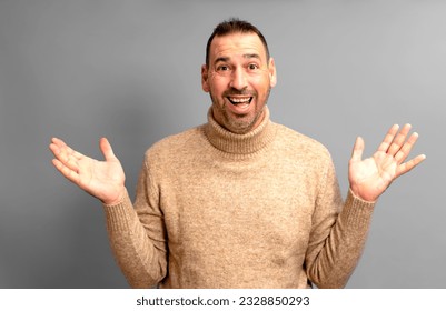 Bearded Hispanic man in his 40s wearing a turtleneck sweater with arms outstretched in an expression of surprise, isolated on gray studio background. - Shutterstock ID 2328850293