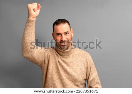 Bearded Hispanic man in his 40s wearing a beige turtleneck raising his fist energetically celebrating a great victory, isolated over gray background