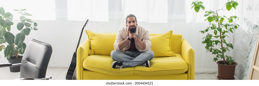 Bearded Hispanic Man Meditating On Yellow Couch In Living Room, Banner