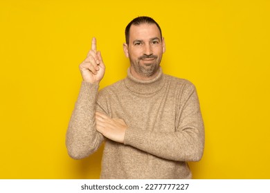Bearded hispanic man wearing a beige turtleneck raising his index finger asking for a turn to express the great idea he had, he feels proud for having found a solution to the problem.