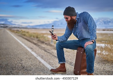 Bearded Hitch Hiker Sitting On Suit Case Using Smart Phone