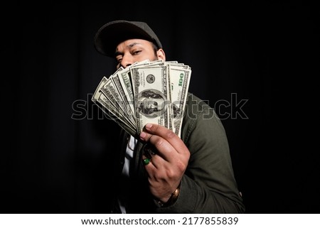 bearded indian hip hop performer in cap covering face with dollar banknotes on black
