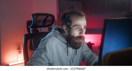 Bearded long-haired hippie looking young adult man playing games at the computer.