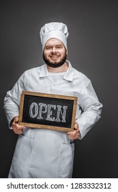 Bearded Male In Chef Uniform Smiling And Looking At Camera While Standing On Gray Background And Holding Blackboard With Open Inscription