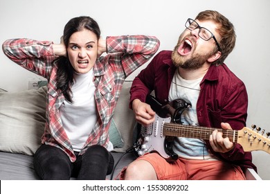 Bearded male composer plays a six-string electric guitar and loudly sings a song of his own composition, his girlfriend does not like this music, she covered ears with hands and bared teeth.
