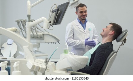 Bearded male dentist and his patient showing their thumbs up at the clinic. Young handsome man sitting on dental chair. Caucasian middle aged doctor in white coat standing near his client