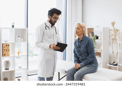 Bearded male doctor and elderly female patient sitting at exam couch of modern medical center and diagnose disease. Young specialist writing instruction for taking medication correctly. - Powered by Shutterstock