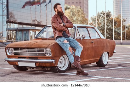 Bearded male dressed in brown leather jacket and boots leaning on tuned retro car in the city parking near skyscraper. - Powered by Shutterstock