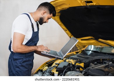 Bearded Male Mechanic Using Laptop, Recording Automobile Engine Checks Collect Detailed Information During His Work On Car Workshop. Service Maintenance During Engine Repair.