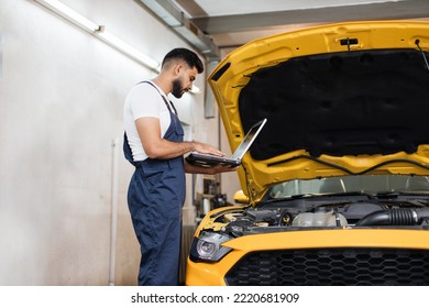 Bearded Male Mechanic Using Laptop, Recording Automobile Engine Checks Collect Detailed Information During His Work On Car Workshop. Service Maintenance During Engine Repair.