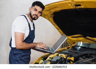 Bearded Male Mechanic Using Laptop, Recording Automobile Engine Checks Collect Detailed Information During His Work On Car Workshop. Service Maintenance During Engine Repair.