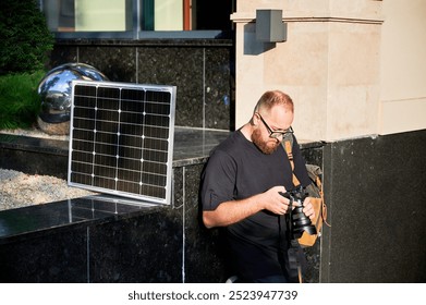 Bearded male photographer reviewing photos on camera, with solar panel set up nearby. Integration of renewable energy in photography. Solar power for eco-friendly charging. - Powered by Shutterstock