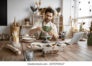 Bearded male seated at wooden table with various tools, materials, and laptop in bright well-decorated room. Smiling man working with clay in cozy home pottery studio. - Powered by Shutterstock