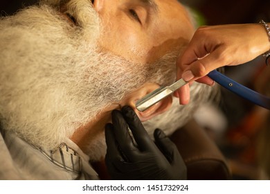 Bearded male sitting in an armchair in a barber shop while hairdresser shaves his beard with a dangerous razor. - Shutterstock ID 1451732924