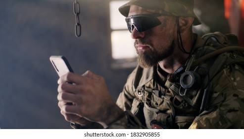 Bearded Male Soldier In Camouflage And Sunglasses Scrolling Data On Smartphone While Resting Inside Abandoned Building During War