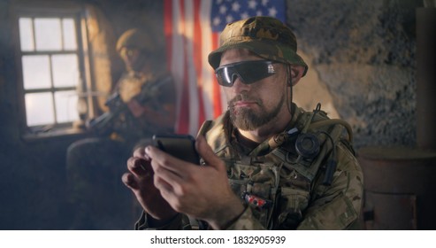 Bearded Male Soldier In Camouflage And Sunglasses Scrolling Data On Smartphone While Resting Inside Abandoned Building During War