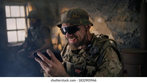 Bearded Male Soldier In Camouflage And Sunglasses Scrolling Data On Smartphone While Resting Inside Abandoned Building During War