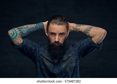 Bearded male with tattooed arms behind his head over dark background.