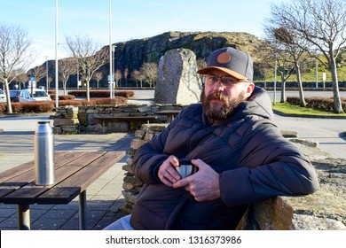 Bearded Man 50 Years Old Drinking A Hot Drink From A Thermos. Norwegian In A Roadside Cafe. Traveling In Norway, Scandinavia, Europe. A Mustached Caucasian Man In Glasses On A Wooden Bench.