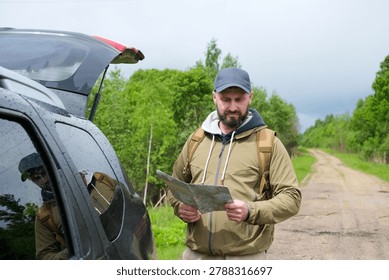 Bearded man with backpack checking paper map beside car with open trunk on country road