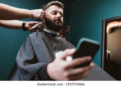 Bearded Man In Barbershop Resting And Looking At His Smart Phone While Hairdresser Gives Him Haircut, Male Beauty Procedures.