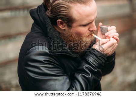 A bearded man in a black leather jacket stands outside and poses for a photographer. Smokes sigaret