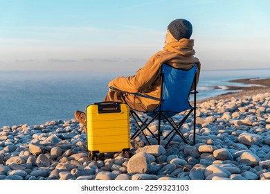 Bearded Man in brown jacket with yellow suitcase relaxing alone and sitting on beach chairs on the seaside at sunrise. Travel Lifestyle concept - Shutterstock ID 2259323103