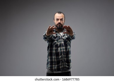 Bearded Man With Calm Face, Wearing Checked Shirt, Standing Against Grey Studio Background And Holding Up A Phone Or Piece Of Glass In His Both Hands Outstretched In Front Of Him