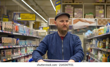 A Bearded Man In A Cap With A Shopping Cart Walks Through The Rows Of Various Goods, Chooses A Purchase For Himself. A Man Is Looking For Goods Jumping From One Shelf To Another. The Man In The Store.