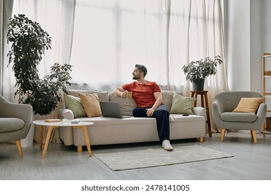 A bearded man in casual attire works remotely from his home office, sitting on a couch with a laptop and a cup of coffee. - Powered by Shutterstock