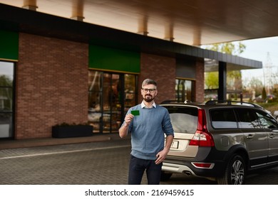 Bearded Man In Casual Clothes Demonstrating Green Plastic Card While Standing Near Car At Gas Station