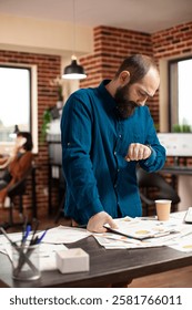 Bearded man checking time and preparing for business project presentation in startup office. Portrait of male analyst looking at his wristwatch and standing at desk filled company research documents.