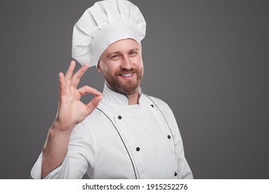 Hombre con barba y uniforme de chef sonriendo para la cámara y mostrando el gesto correcto mientras aprueban el servicio de restaurante con fondo gris