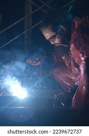 A bearded man construction worker doing welding activity at night in factory. Metal work industrial concept.