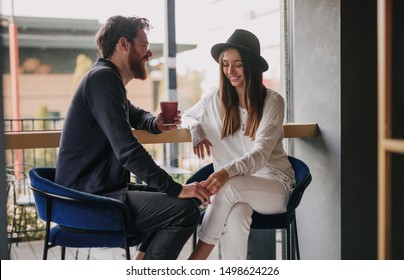 Bearded man with cup of hot drink smiling and touching hand of beautiful young woman while sitting near window in cozy cafe and flirting, during first date
