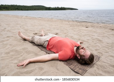 Bearded Man Does Yoga, He Does The Savasana Exercise, Relaxes In A Corpse Posture Lying On A Rug, On A Sandy Beach By The Lake.