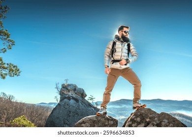A bearded man in a down jacket and backpack hikes a rocky peak with mountains in the background and the sun shining behind him.