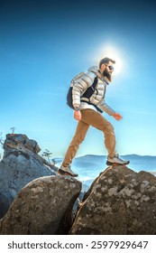 A bearded man in a down jacket and backpack hikes a rocky peak with mountains in the background and the sun shining behind him.