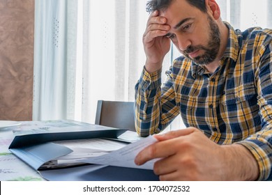 Bearded man dressed in plaid shirt looking at bills worried in his living room. Despair concept - Shutterstock ID 1704072532