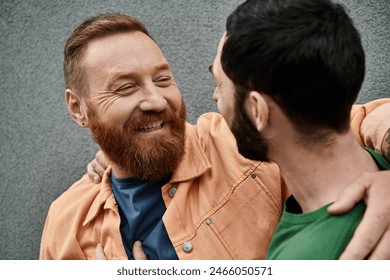 A bearded man embraces another man lovingly against a grey wall, both in casual attire.