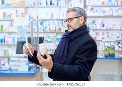 Bearded Man Frowning, Holding White Bottle Mock And Prescription Paper In Hands. Customer Wearing Eyeglasses And Scarf. Old Man Reading, Choosing Medical Products In Apothecary Store.